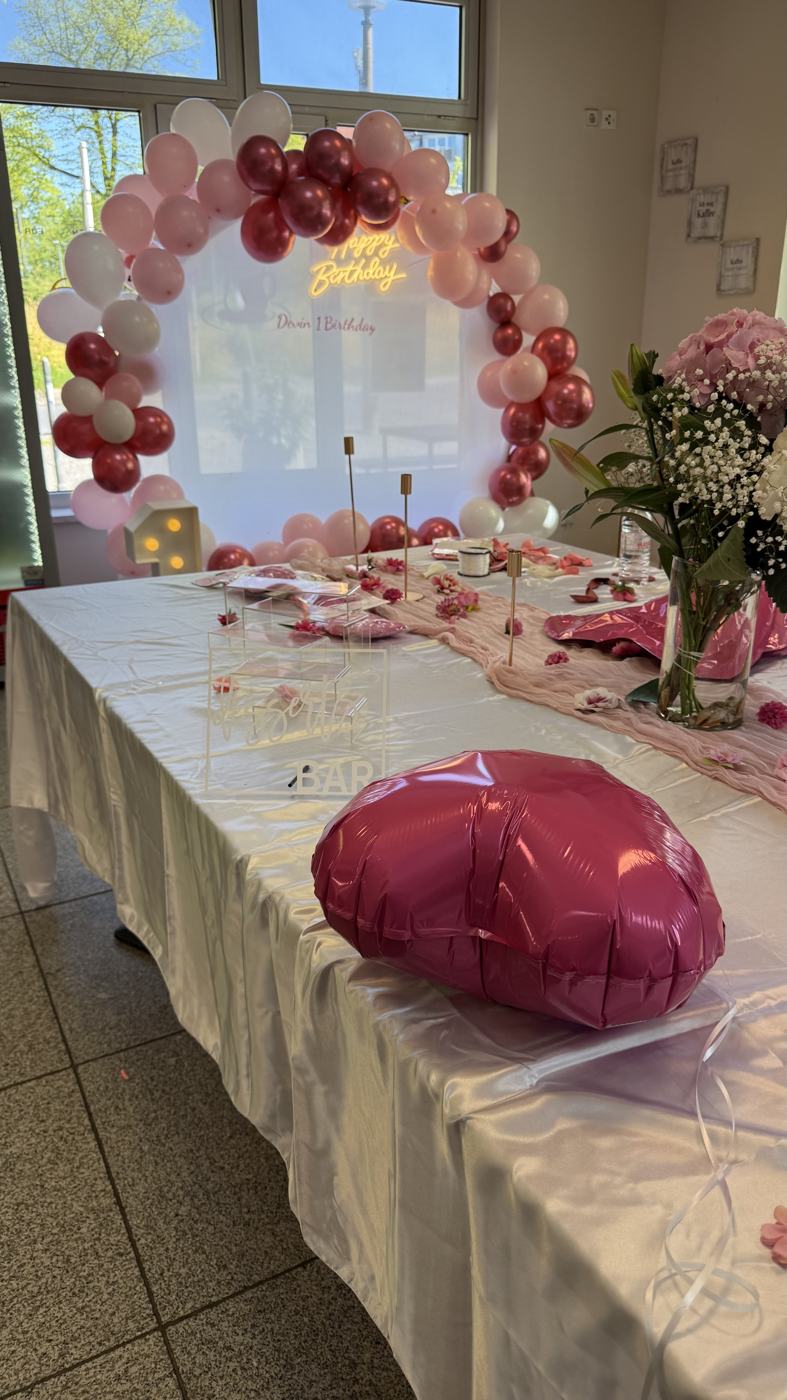 Rosa dekoriertes Geburtstagsbuffet mit Ballonbogen, Neon-Schriftzug "Happy Birthday" und Blumendekoration